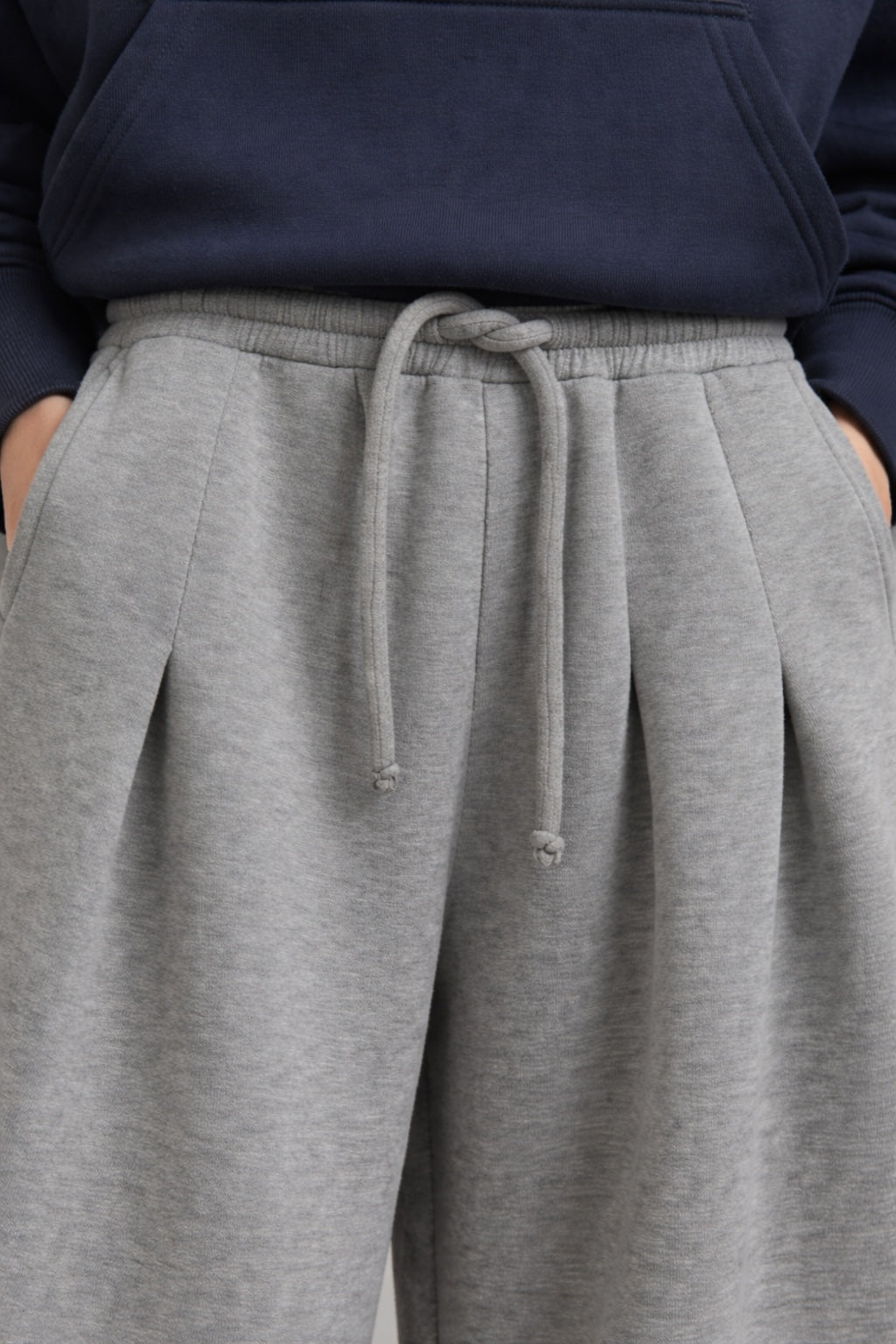 Close-up of a person wearing heather gray pleated sweatpants with a matching drawstring waist, paired with a navy hoodie.